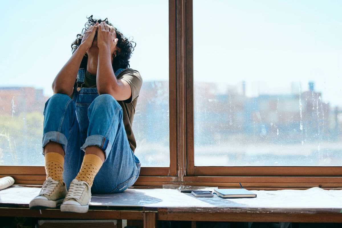 Erschöpfte junge Frau sitzt auf einem Fensterbrett.